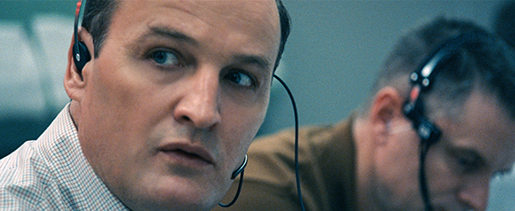 A man in a checkered shirt wearing a headset looks to the side with a serious expression.