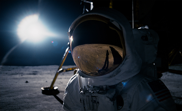 An astronaut in a white spacesuit with a reflective gold visor stands on the moon's surface near a bright light.