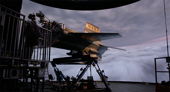 A NASA aircraft model mounted on a motion platform in front of a large screen showing clouds.