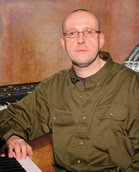 A man with glasses and an olive green button-down shirt sits behind a musical keyboard.