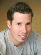 A man with short dark hair and a light beard smiling while wearing a grey t-shirt.