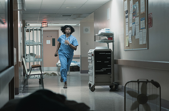 A nurse in blue scrubs runs down a hospital corridor past medical carts and equipment.