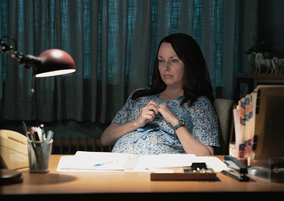 A pregnant woman in a floral dress sits at a desk with papers and a lamp in a dimly lit room.