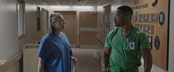 A man in green scrubs talks to a woman in blue scrubs while walking down a hospital corridor.