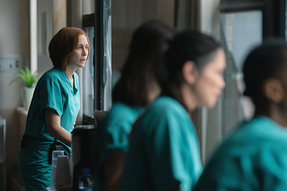 A woman in teal scrubs stares intently out a window while others in similar scrubs stand in the foreground.