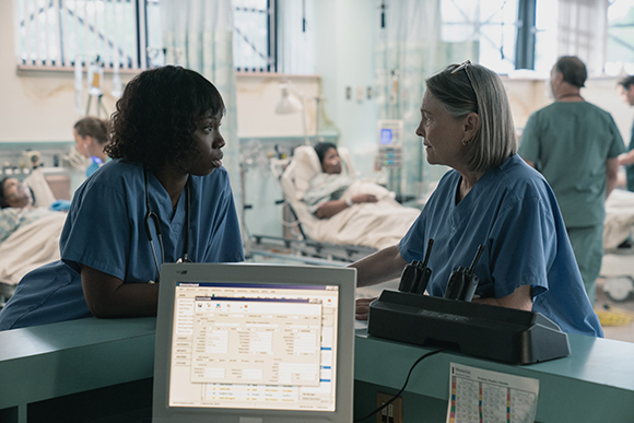 Two nurses in blue scrubs converse behind a hospital desk with a computer monitor in the foreground.
