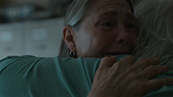 An elderly woman with grey hair cries while embracing another person in a close-up shot.