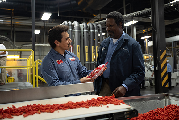 Two men in blue work shirts stand in a factory near a conveyor belt filled with small red food items.