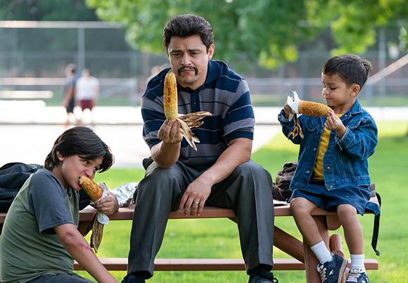 A man sits on a wooden bench between two young boys, all of whom are eating corn on the cob.