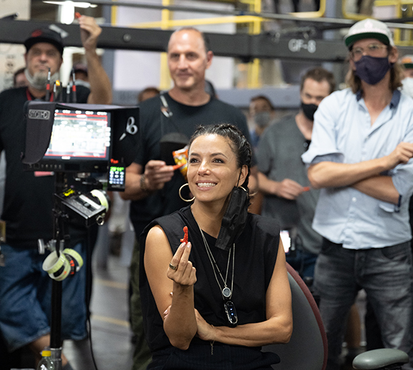 A woman in a black sleeveless top smiles while sitting on a film set with crew members in the background.