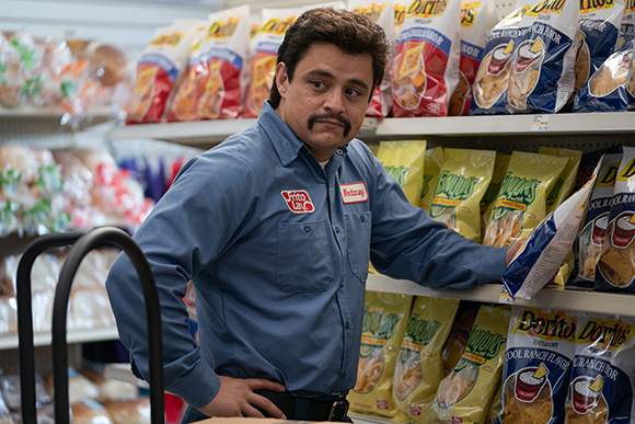 A man in a blue button-down shirt stands in a grocery aisle next to shelves of snack bags.