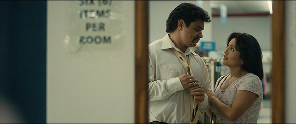 A man in a white shirt and a woman in a patterned top look at each other through a mirror reflection.