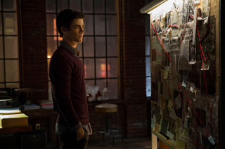 A man in a maroon sweater stands in a dimly lit room looking at a corkboard covered in notes and red string.