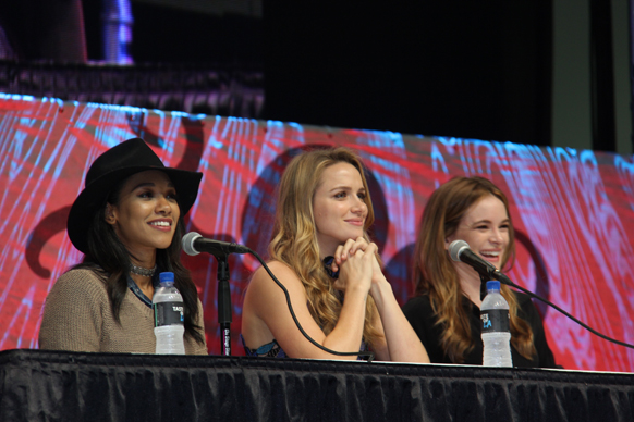 Three women sit at a panel table with microphones and water bottles. The woman on the left wears a black hat and a tan sweate