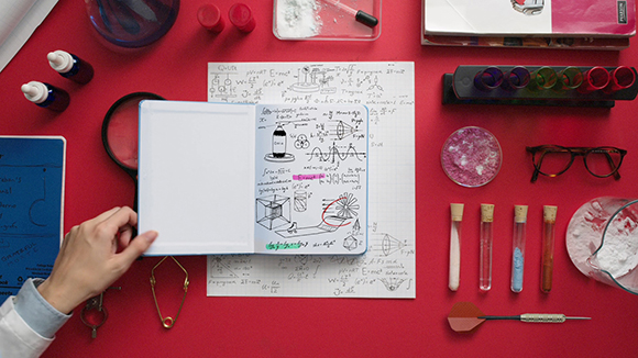 Top-down view of a red desk with a notebook, scientific glassware, test tubes, and physics diagrams.