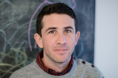 A man with dark hair and green eyes wearing a grey sweater over a red plaid shirt.
