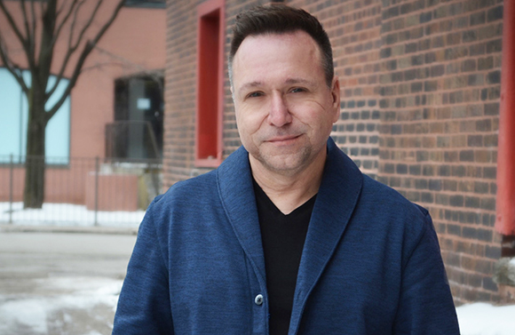 A man with short dark hair wearing a blue jacket and black shirt stands before a brick wall.