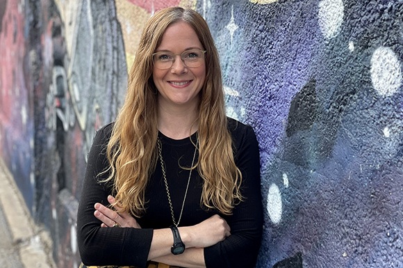 A smiling woman with long wavy hair and glasses stands with arms crossed against a purple and blue mural.