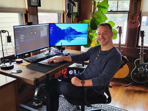 A smiling man sits at a wooden desk with two computer monitors, a keyboard, and musical instruments nearby.