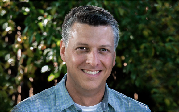 A smiling man with short salt-and-pepper hair wearing a blue plaid shirt against a leafy green background.