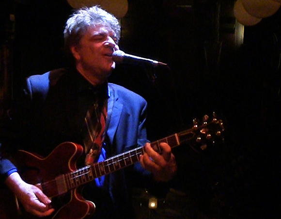 A man in a suit playing an electric guitar and singing into a microphone under dim lighting.