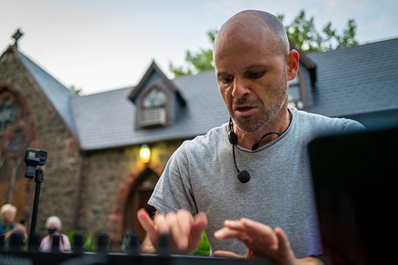 A bald man in a grey t-shirt plays a keyboard with a headset microphone on.