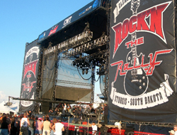A large outdoor concert stage with black banners reading Rock'n the Rally, Sturgis, South Dakota.