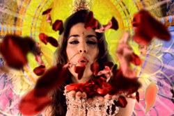 A woman blowing red rose petals toward the camera against a bright, circular patterned background.