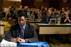 A man in a dark suit sits with hands clasped at a table, facing a large audience in a conference setting.
