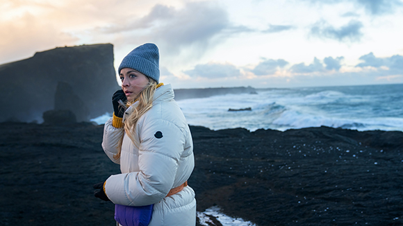 A woman in a white puffer jacket and blue beanie looks out over a dark, rocky coastline with crashing waves.