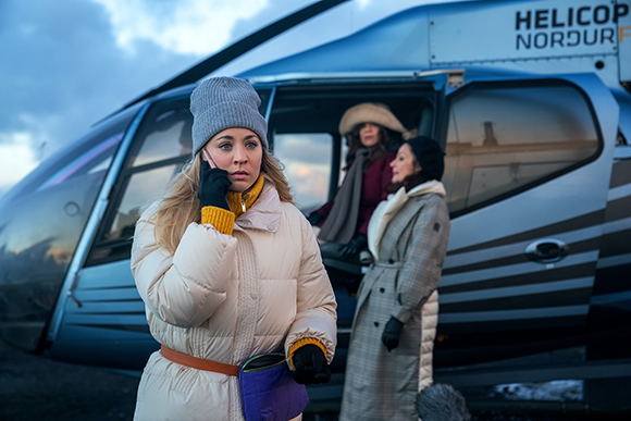 A woman in a white puffer jacket and grey beanie talks on a phone next to a blue helicopter.