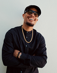 A smiling man wearing sunglasses, a black baseball cap, and a gold chain leans against a white wall.