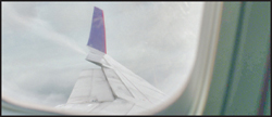 An airplane wing with a purple vertical stabilizer seen through a passenger window against a cloudy sky.
