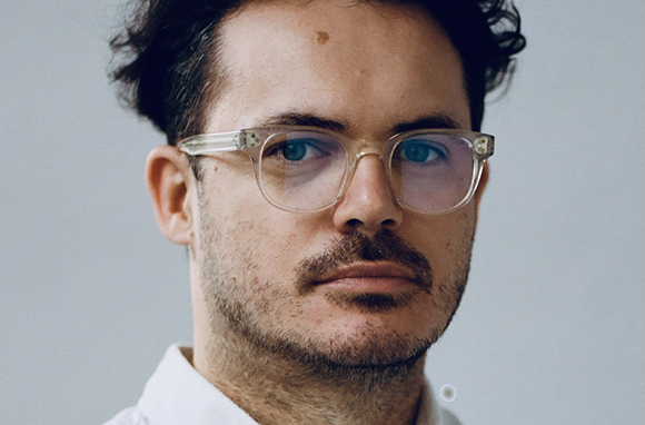 Close-up portrait of a man with dark hair and clear-framed glasses looking forward.