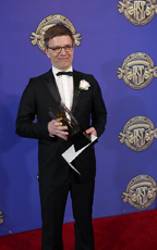 A man in a black tuxedo and glasses holds a trophy and a white paper against a blue backdrop.