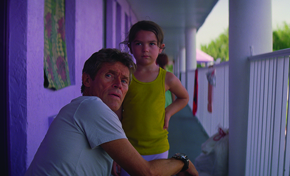 A man in a white shirt and a young girl in a yellow tank top stand on a purple porch.