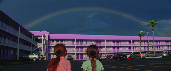 Two children stand in a parking lot looking at a large rainbow arching over a two-story purple motel building.