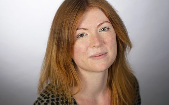 Close-up portrait of a woman with long red hair and light eyes looking toward the camera.