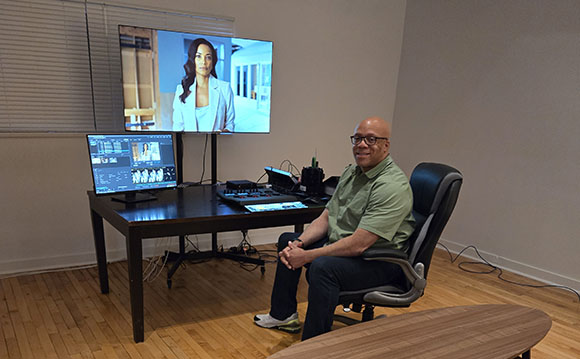 A man in a green shirt sits in an office chair before a desk with monitors and video production equipment.