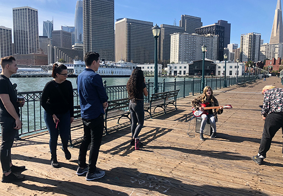 People stand on a wooden boardwalk near a waterfront with city skyscrapers in the background.