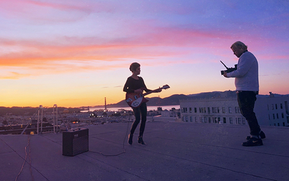 A person plays guitar on a rooftop at sunset while another person films them with a camera.