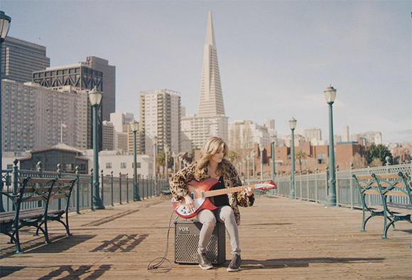A woman sits on a boardwalk playing a red electric guitar with a city skyline and Transamerica Pyramid behind her.