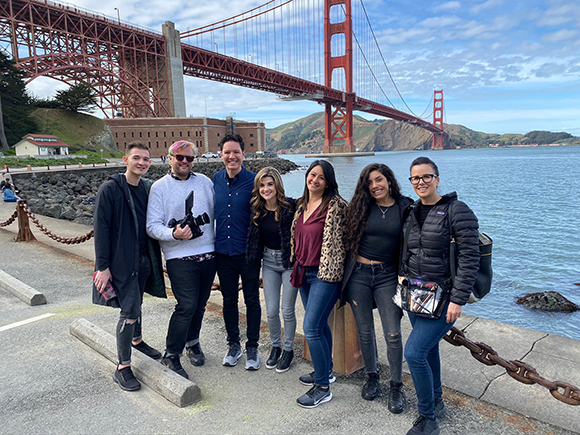 Seven young adults standing in a row on a paved walkway with the Golden Gate Bridge in the background.