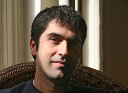 A man with short dark hair and dark eyes looking forward while sitting in a wicker chair.