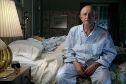 An elderly man in light blue pajamas sits on a bed next to a person lying in bed under a lamp.