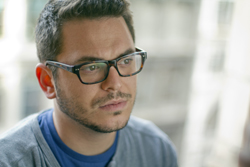A man with dark hair and glasses looking to the side against a blurred background.