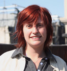 A smiling woman with shoulder-length red hair wearing a grey shirt and white jacket.