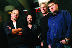 Four men standing together in a dimly lit indoor setting.