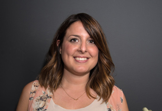 A smiling woman with shoulder-length brown hair wearing a floral sleeveless top against a dark background.