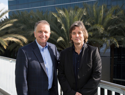 Two men in blazers standing side by side in front of palm trees and a glass building.